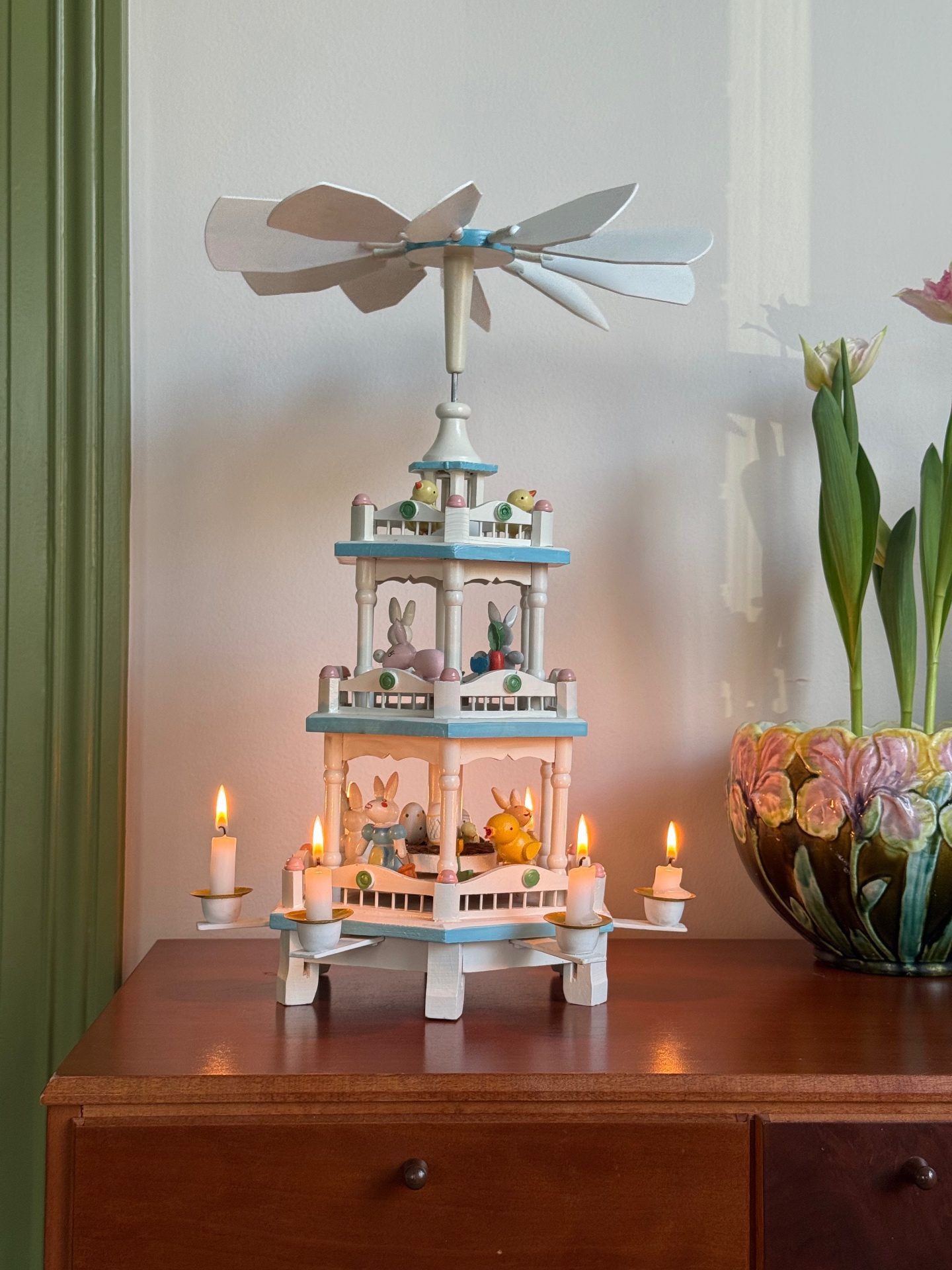 Painted Easter pyramid with rabbits and lit candles on a sideboard beside tulips