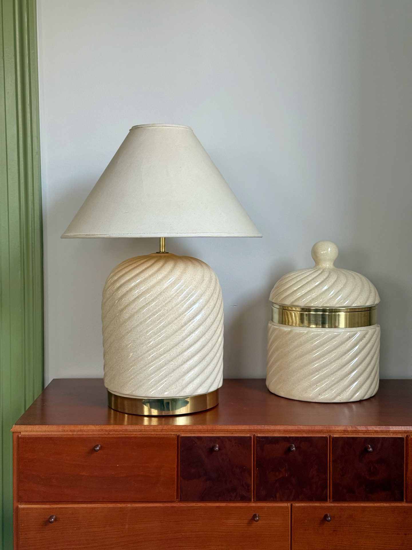 Cream ceramic ice bucket with a brass rim beside a matching table lamp on a wooden sideboard